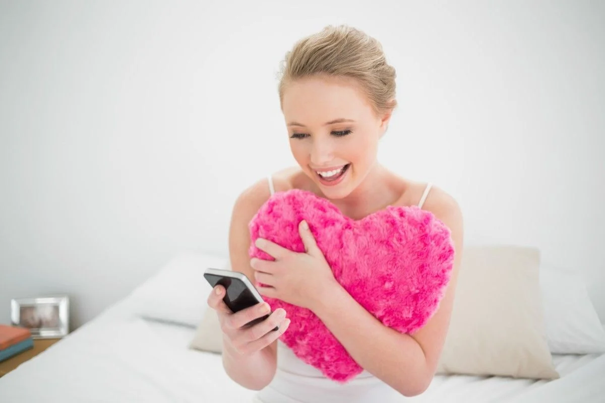 A woman with a heart-shaped pillow smiling at her phone in a cozy bedroom. Chat Up Lines for Ladies That Are Playful and Romantic