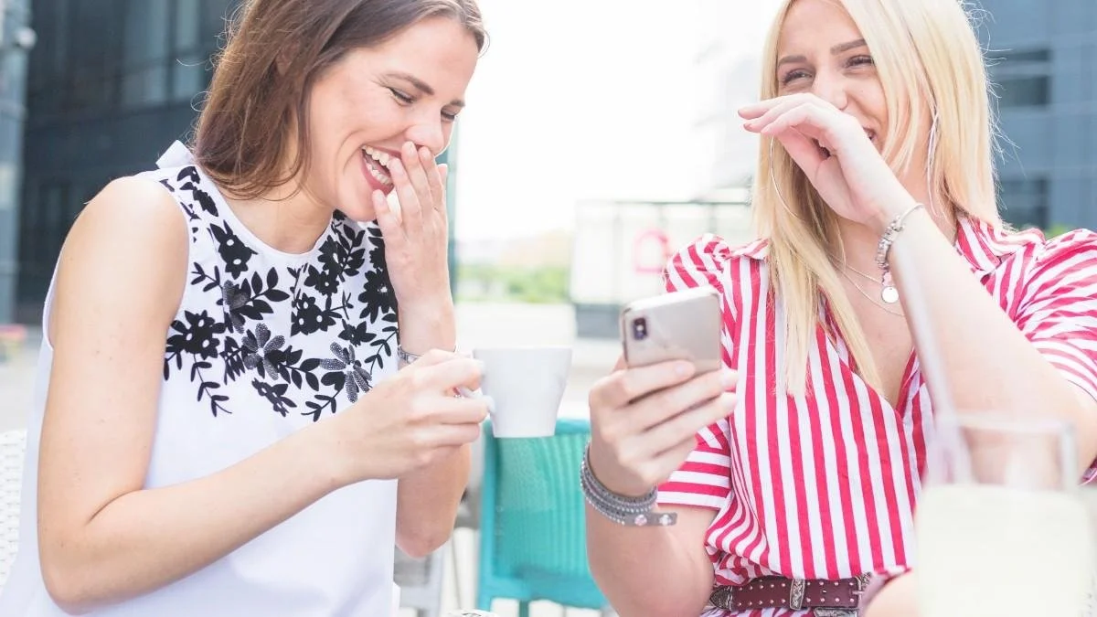 Two smiling women enjoying a laugh at an outdoor cafe while one holds a cup of coffee and the other checks her phone.Chat Up Lines for Ladies That Are Playful and Romantic.